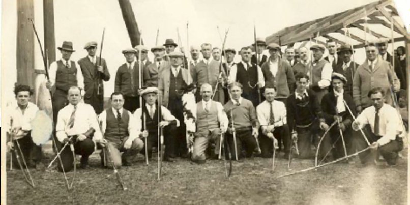 Early club members standing and kneeling with their bows and poles in front of a shed